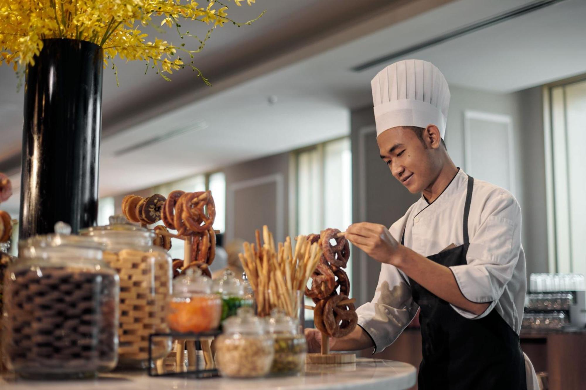 מלון Jw Marriott קואלה לומפור מראה חיצוני תמונה The photo shows a young chef in a professional kitchen setting. He is wearing a white chef's hat and a dark apron over a white chef's jacket. The chef is arranging pretzels on a table or display. In the background, there are several jars filled with 