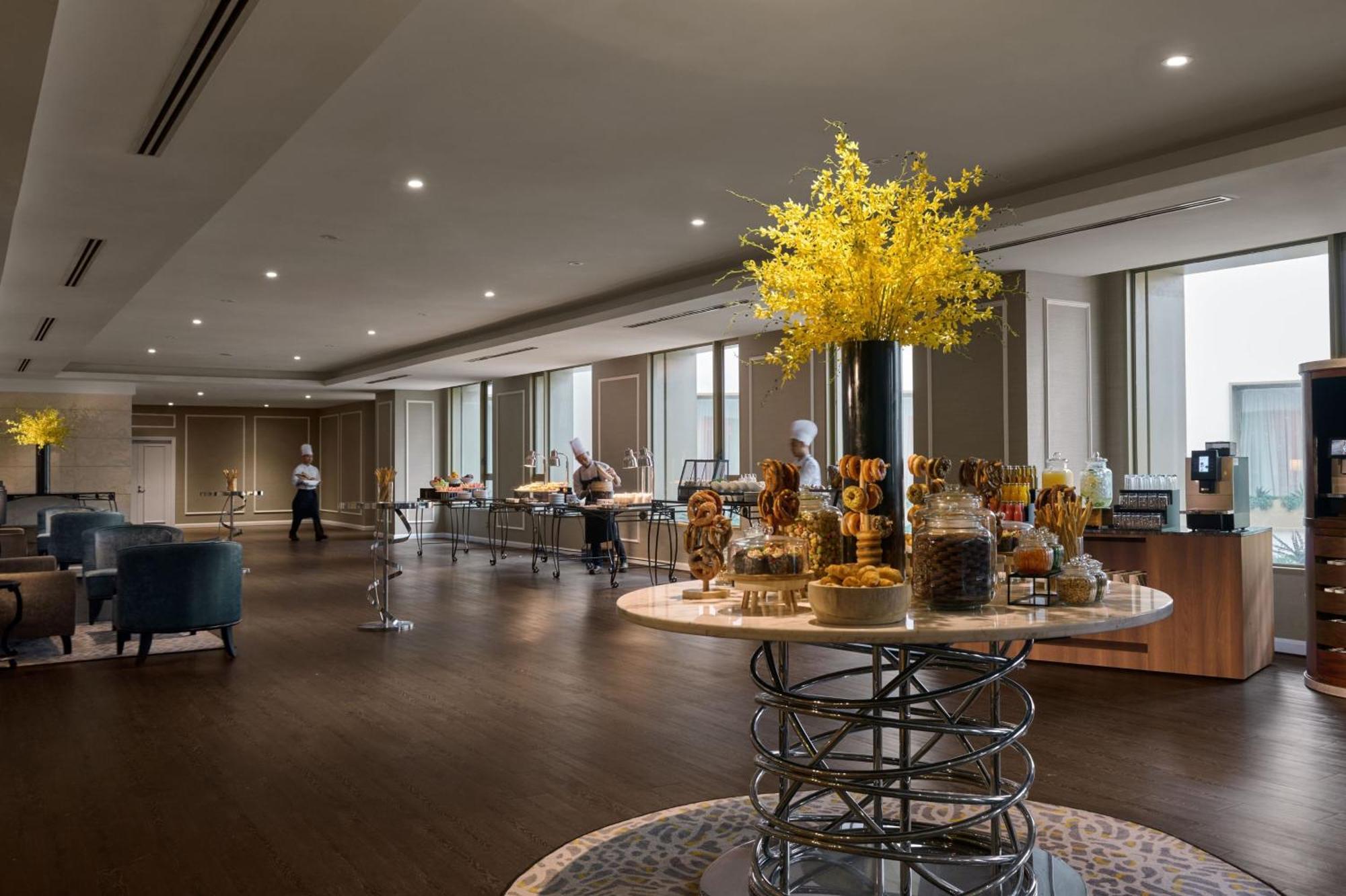 מלון Jw Marriott קואלה לומפור מראה חיצוני תמונה The photo shows a modern, elegant dining area, likely set up for a buffet or catering event. In the center, there is a large, round table with a decorative arrangement of yellow flowers, accompanied by various jars and displays of food items. Several